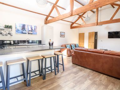 An open-plan living room and kitchen with wooden beams a countertop with stools leather sofas and a wall-mounted TV at The Barnhouse in Felmingham near North Walsham