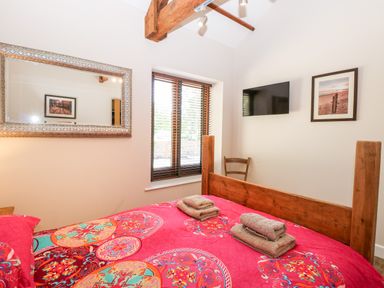 A bedroom with a wooden bedframe and red patterned bedspread a window with blinds a wall-mounted television a mirror and framed pictures at The Barnhouse in Felmingham near North Walsham