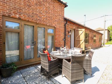 An outdoor patio with a round glass table set with plates and glasses surrounded by six wicker chairs in front of a brick building at The Barnhouse in Felmingham near North Walsham