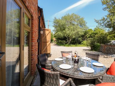 An outdoor dining area with a glass table set with plates and glasses and wicker chairs by a brick building at The Barnhouse in Felmingham near North Walsham