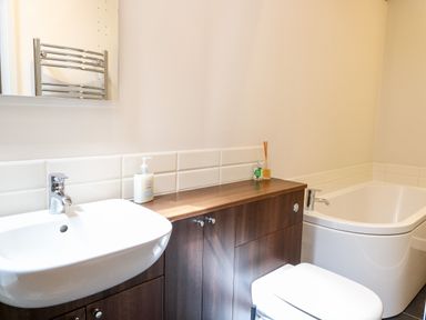 A bathroom with a white sink a wooden cabinet a bathtub a toilet and a wall-mounted mirror at The Barnhouse in Felmingham near North Walsham