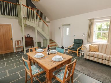 A dining and living area with a wooden table set for four chairs a sofa armchair a staircase and a window at Ystabl Robyn in Llanddarog