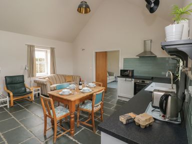 A kitchen and dining area with wooden table and chairs dishes and wine bottle a beige striped sofa a green armchair and kitchen appliances at Ystabl Robyn in Llanddarog