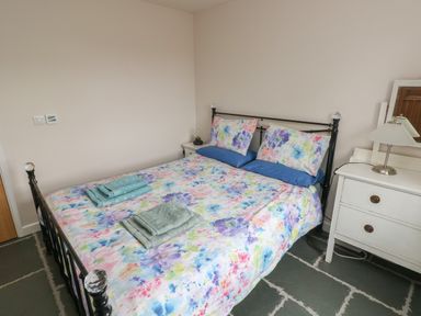 A bedroom with a double bed with floral bedding blue pillows towels on bed a white bedside table with lamp and mirror at Ystabl Robyn in Llanddarog