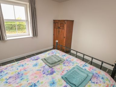 A bedroom with a floral bedspread two folded towels a window with curtains and a wooden wardrobe at Ystabl Robyn in Llanddarog