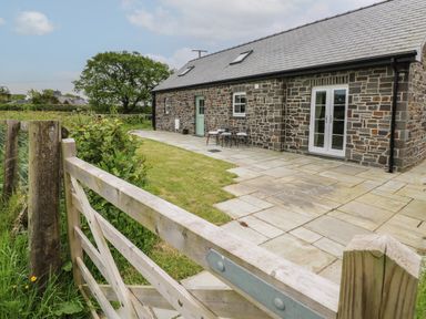 A stone house with a slate roof a wooden gate a small lawn and a paved patio with outdoor chairs at Ystabl Robyn in Llanddarog