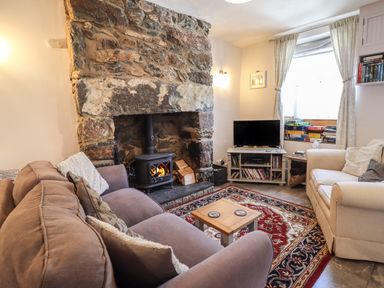 A living room with a stone fireplace a wood stove a beige armchair a brown sofa a wooden coffee table a red patterned rug a tv on a stand and board games by the window at Llygoden Cottage in Beddgelert