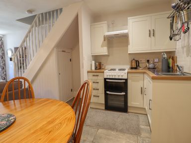 A kitchen with white cabinets an oven a wooden dining table and chairs under stairs at Llygoden Cottage in Beddgelert