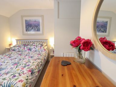 A bedroom with a floral bedspread a wooden dresser with flowers and a round mirror at Llygoden Cottage in Beddgelert