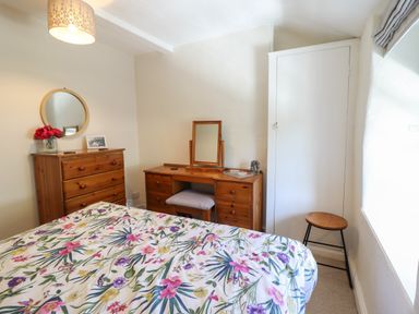 A bedroom with floral bedspread a wooden dresser with mirror and flowers a wooden dressing table with stool a small wooden stool and white closet door at Llygoden Cottage in Beddgelert
