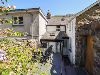 An outdoor patio area with black metal table and chairs next to a stone wall and a white house with a small door at Llygoden Cottage in Beddgelert