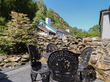 An outdoor seating area with a black metal table and four chairs next to a stone wall and houses at Llygoden Cottage in Beddgelert