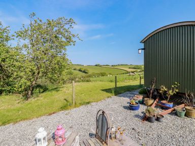 Outdoor seating area with lanterns and potted plants next to a green metal hut overlooking green fields at Puddle Duck Shepherds Hut in Crackington Haven