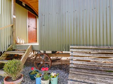 An exterior view of a corrugated metal shed with wooden steps and a red door with potted plants and a weathered wooden bench near the entrance at Puddle Duck Shepherds Hut in Crackington Haven