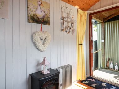 An interior corner with a small stove, a heater, a painting of a duck, a heart-shaped wreath, hanging decorations, yellow curtains, and an open door showing a porch with duck figurines at Puddle Duck Shepherds Hut Crackington Haven