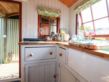 A small kitchen corner with cabinets a window with floral blinds and kitchen items on the counter at Puddle Duck Shepherds Hut in Crackington Haven