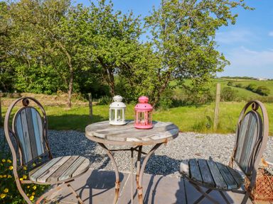 An outdoor patio area with a round wooden table and two chairs with lanterns on the table and greenery in the background at Puddle Duck Shepherds Hut in Crackington Haven