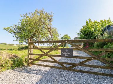 A wooden gate with a Puddle Duck Hut sign and a gravel driveway with trees on each side at Puddle Duck Shepherds Hut in Crackington Haven
