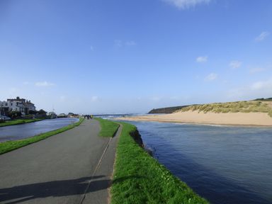 A paved path with grass on both sides running alongside a river and a sandy beach with dunes at Puddle Duck Shepherds Hut in Crackington Haven