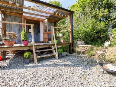 An outdoor area with a wooden porch, potted plants, small metal table and chairs, and a fire pit at Shepherds Retreat in Maxworthy near Crackington Haven