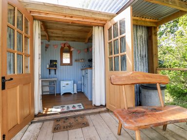 A wooden bench outside open double doors leading to a small room with a light blue stove and kitchen area at Shepherds Retreat Maxworthy near Crackington Haven