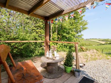 A covered wooden porch with a wooden chair and spool table with lanterns and flowers overlooking a rural landscape at Shepherds Retreat in Maxworthy near Crackington Haven