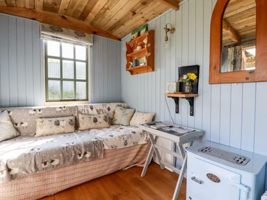 A small room with a wooden ceiling a window a sofa with sheep patterned covers a small table a wall shelf with decorations a mirror and a blue stove at Shepherds Retreat Maxworthy near Crackington Haven
