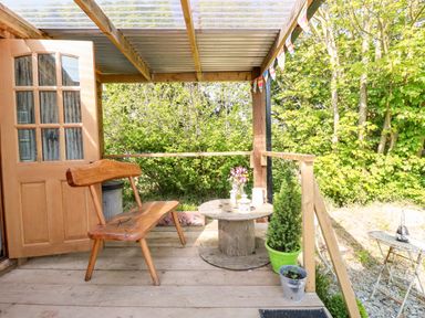 A wooden porch with a bench wooden round table potted plants and an open door at Shepherds Retreat Maxworthy near Crackington Haven