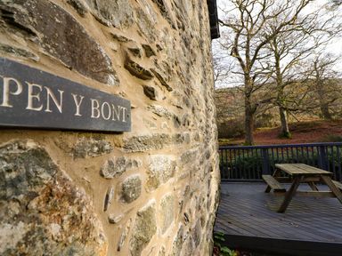A stone wall with a sign reading Pen y Bont and a wooden deck with a picnic table near trees and hills at Pen y Bont Near Mallwyd