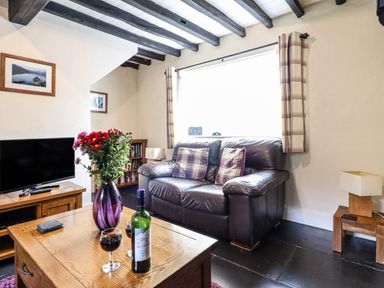 A living room with a brown leather sofa and two plaid pillows a wooden coffee table with a purple vase of flowers a bottle of wine two glasses a television on a wooden stand two framed pictures on the wall and a wooden side table with a lamp at Pen y Bont Near Mallwyd