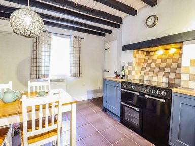 A kitchen with a wooden dining table and chairs next to a black stove with blue cabinets and a window with checkered curtains at Pen y Bont Near Mallwyd