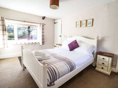 A bedroom with a white wooden bed a purple cushion and striped bedding a nightstand and a window with curtains at Pen y Bont Near Mallwyd