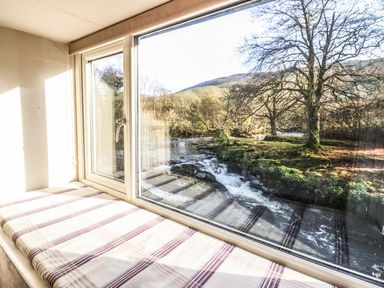 A window with a cushioned seat overlooking a river and trees at Pen y Bont Near Mallwyd