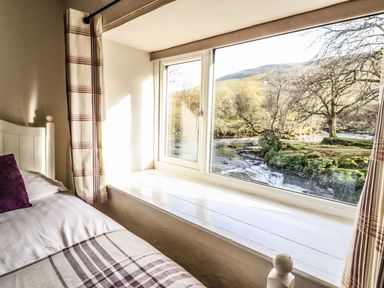 A bedroom window with plaid curtains and a bed next to it showing a river and trees outside at Pen y Bont Near Mallwyd