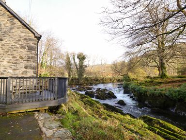 A stone house with a wooden deck next to a river with rocks and trees near Pen y Bont Near Mallwyd