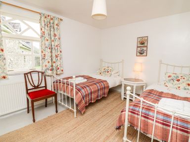 A bedroom with two single iron beds with blankets and pillows a wooden chair and a window with floral curtains at Heron House in Talley near Llandeilo