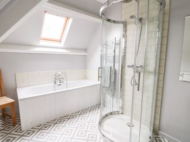 A bathroom with a bathtub under a skylight window a shower cubicle with glass doors and a wooden chair on patterned floor tiles at Heron House in Talley near Llandeilo