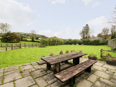 A garden with a wooden picnic table on a stone patio and a grassy field with trees and hills at Buzzards Watch in Talley near Llandeilo