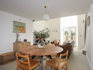 A dining area with a round wooden table and chairs next to a living room with sofas and large windows at Ysgol Fach (Little School) in Benllech
