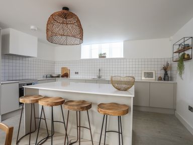 A kitchen with a white island and four wooden stools under a wicker pendant light at Ysgol Fach Little School in Benllech