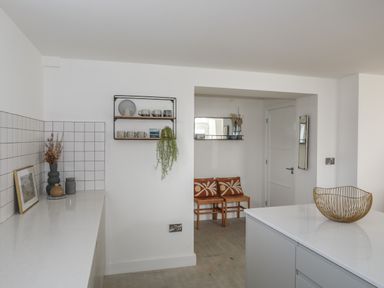 A kitchen with white countertops a shelf with mugs a decorative bowl and two chairs with cushions at Ysgol Fach Little School in Benllech