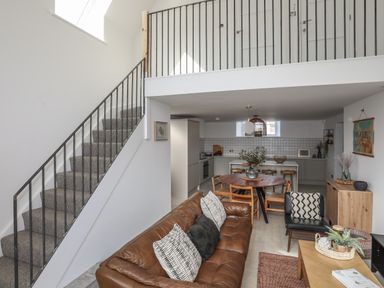 An open-plan living area with a brown leather sofa and black armchair facing a wooden coffee table dining table with chairs and a kitchen in the background with stairs leading to an upper level at Ysgol Fach Little School in Benllech