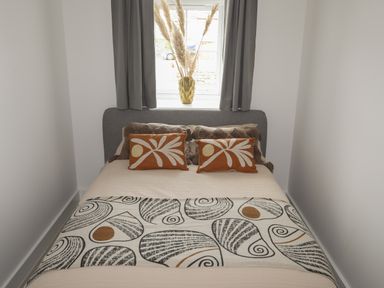 A bedroom with a bed and decorative pillows under a window with curtains and a vase with dried plants at Ysgol Fach Little School in Benllech