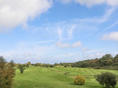 A golf course with green grass fairways and scattered trees under a blue sky at Ysgol Fach Little School in Benllech