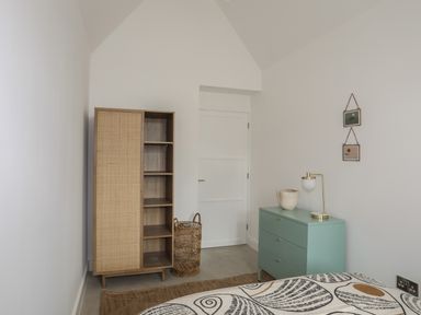 A bedroom with a wooden cabinet a wicker basket a green chest of drawers with a lamp and vase and two small framed pictures on the wall at Ysgol Fach Little School in Benllech