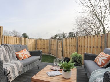 An outdoor patio with two gray sofas, a wooden table, plants, and a wooden fence at Ysgol Fach Little School in Benllech