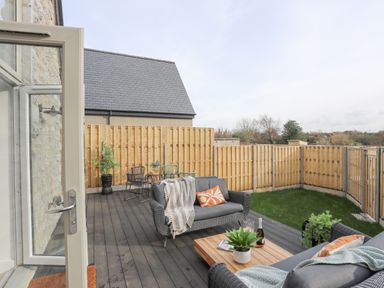 An outdoor patio with wicker sofas and a wooden table with plants and a bottle next to a grassy yard and wooden fence at Ysgol Fach Little School in Benllech