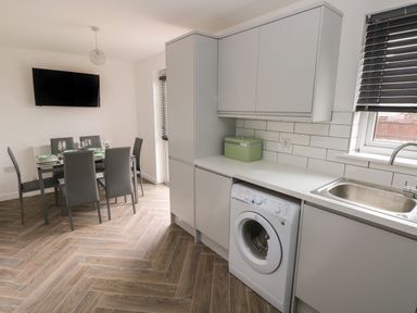 A kitchen with a washing machine under the counter a sink window with blinds and a dining table with six chairs and a wall mounted television at 39 Mariners Quay in Port Talbot
