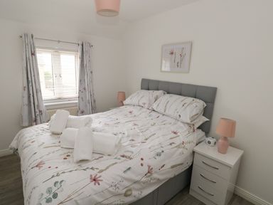 A bedroom with a double bed with floral bedding beside a window with curtains and a white bedside table with a pink lamp and an alarm clock at 39 Mariners Quay in Port Talbot