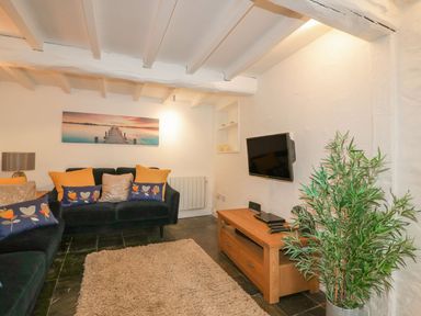 A living room with black sofas yellow and floral cushions a wall-mounted TV and a potted plant at Blacksmiths Cottage in Crantock